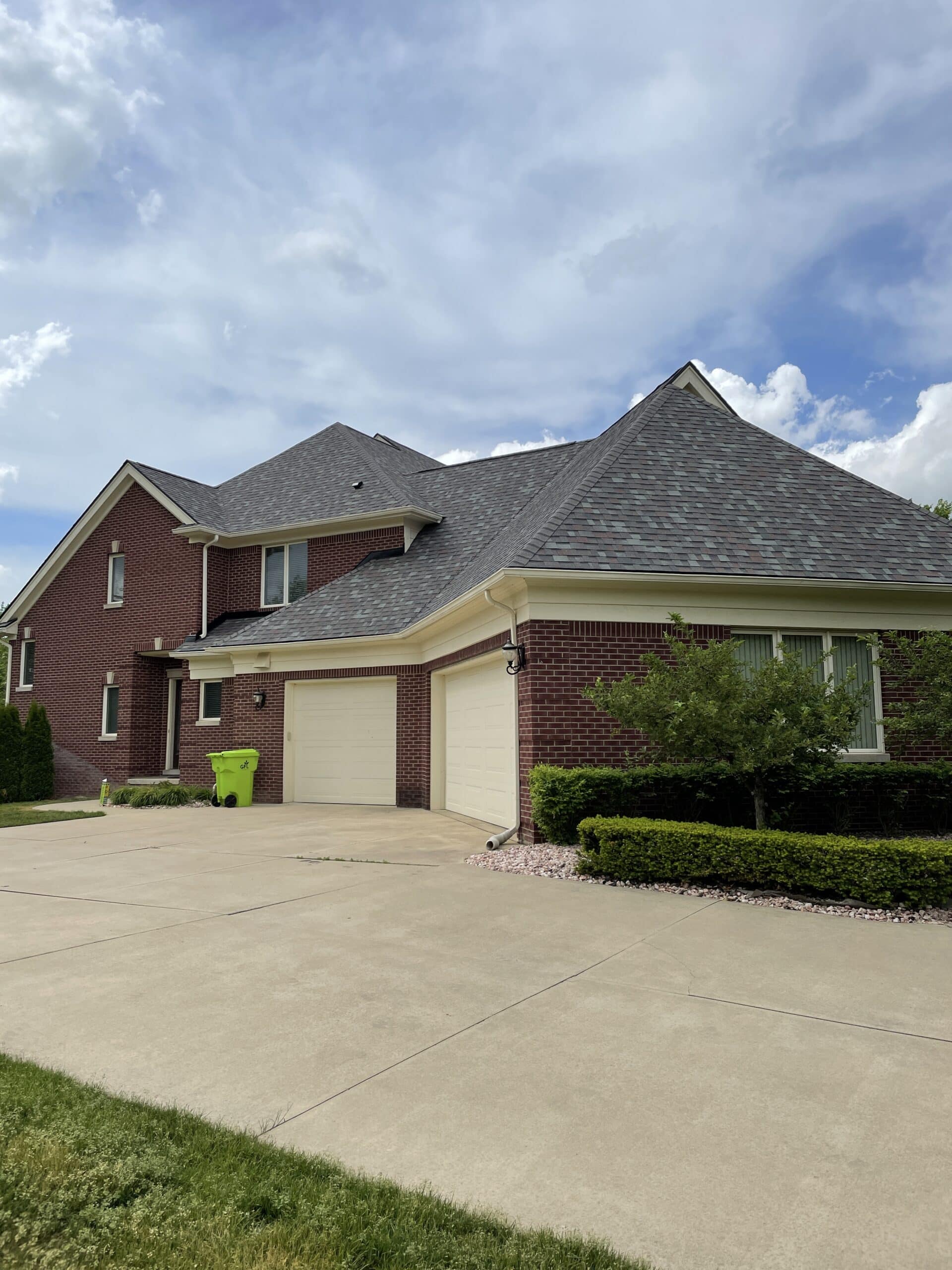 A large brick house with a steeply sloped shingle roof and a three-car garage, under a partly cloudy sky.