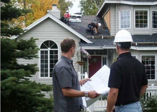 Two men on the ground discussing over a blueprint, while another man on a house roof performs maintenance, suburban setting.
