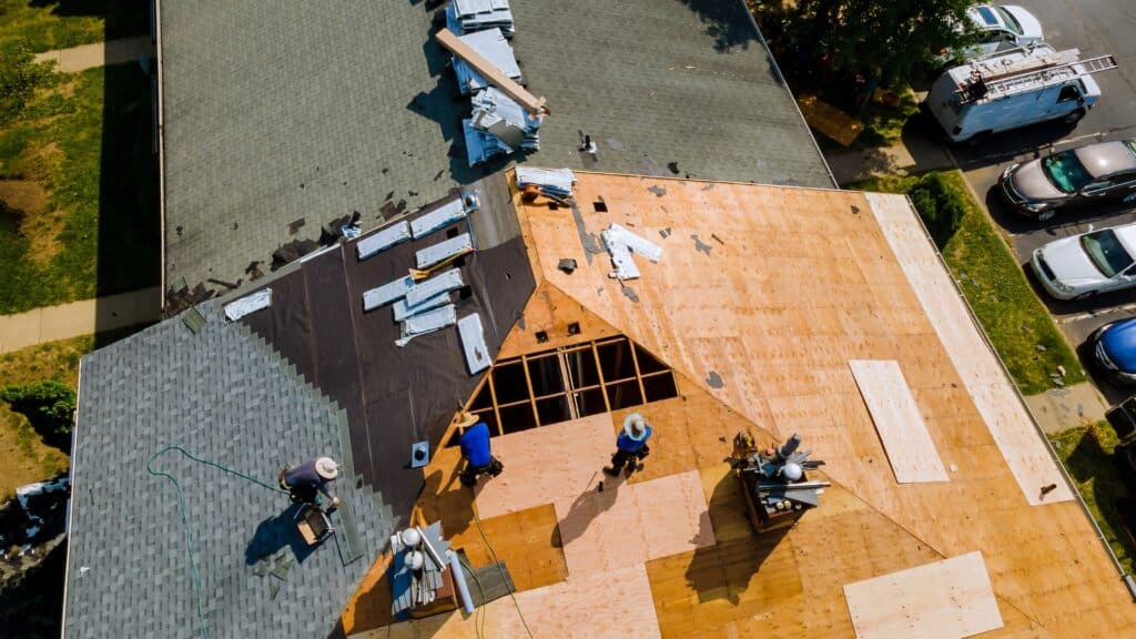 Aerial view of roofers working on a house, with part of the roof covered in new plywood and another section maintained with green shingles. Several tools and materials are scattered around.