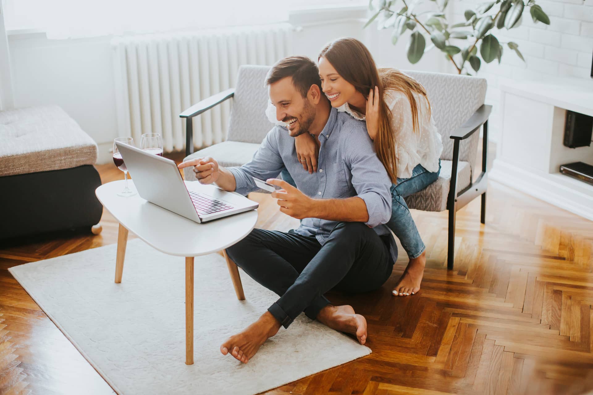 A couple sits on the floor in a living room, smiling and looking at a laptop on a coffee table.