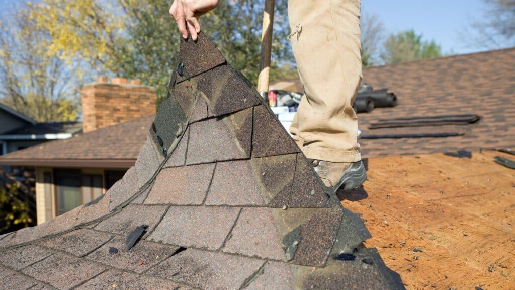 A person is removing worn shingles from the roof of a house, exposing the wooden structure beneath. Various tools and materials are visible in the background.