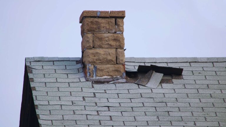 A brick chimney on a shingled roof shows visible roof damage