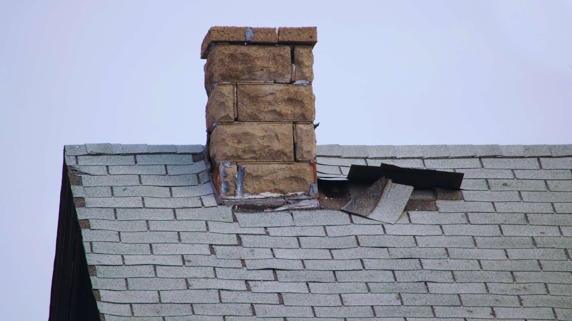 A brick chimney on a shingled roof shows visible roof damage