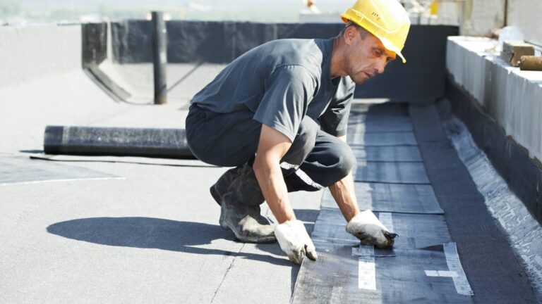A construction worker wearing a yellow hard hat and gloves performs roof repair, carefully installing roofing material on a flat rooftop.