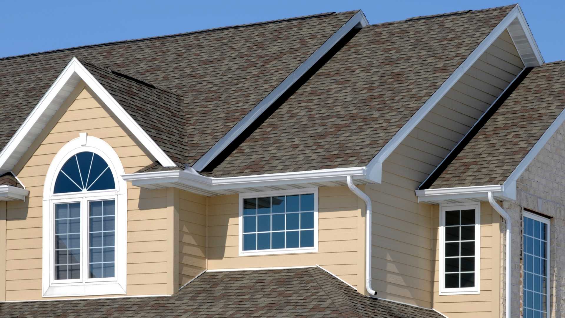 The image shows the exterior of a house with beige siding, white trim, large windows, and a dark shingle roof under a clear blue sky.
