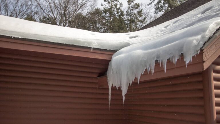 Large icicles and a thick layer of snow hang from the edge of a brown house roof on a cold winter day.