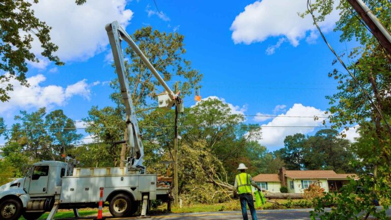 utility workers use a bucket truck to repair power lines after a fallen tree caused damage near a residential area.